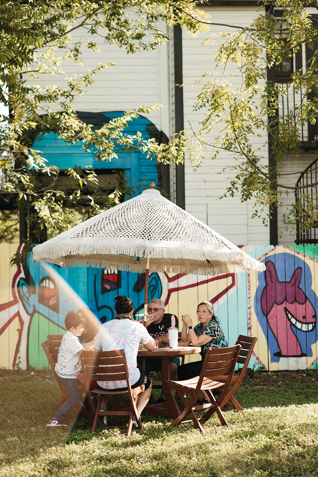 People at a wooden table under a fringed umbrella in front of a painted mural fence