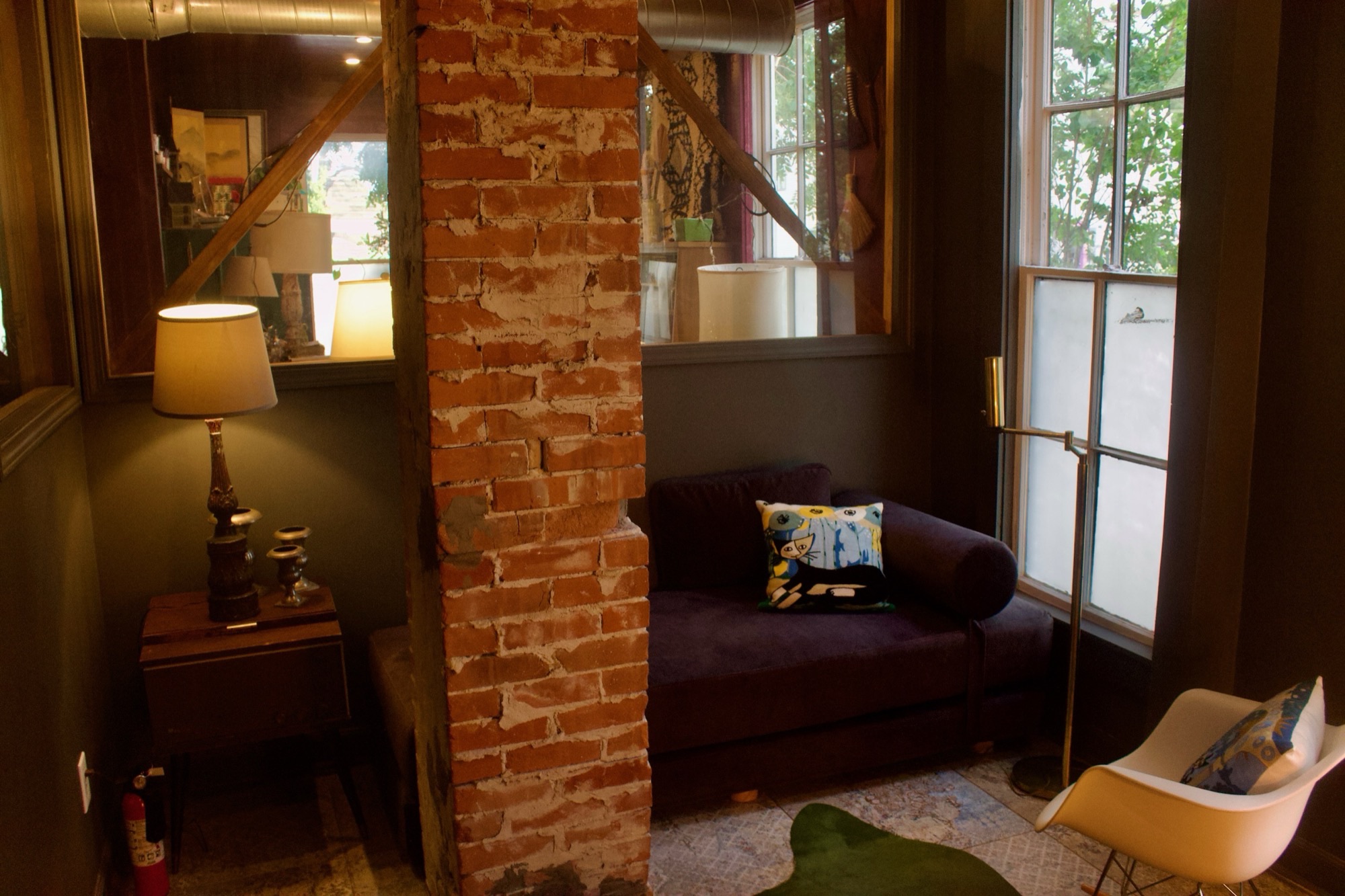 A studio room with a brick column, dark sofa, lamp and window